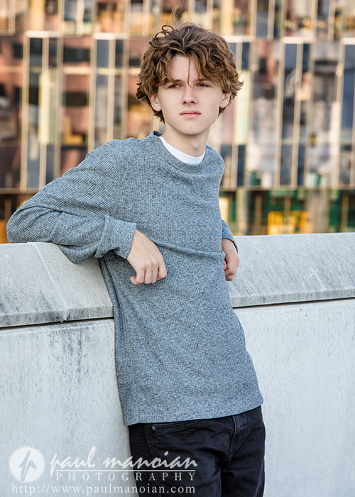 A young person with wavy brown hair, wearing a gray sweater and black pants, leans against a concrete ledge during a Detroit senior pictures session, with a modern glass building in the background.