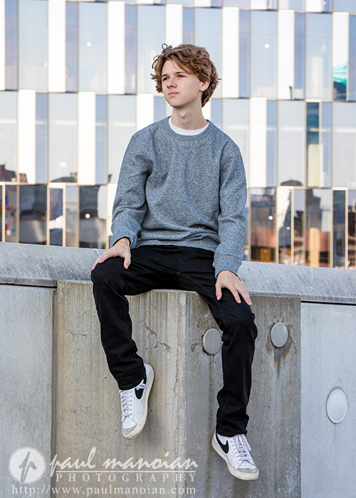 A teenage boy with curly hair, wearing a gray sweatshirt, black pants, and white sneakers, sits on a concrete ledge outdoors in front of a modern building—perfect for a Detroit senior pictures session.