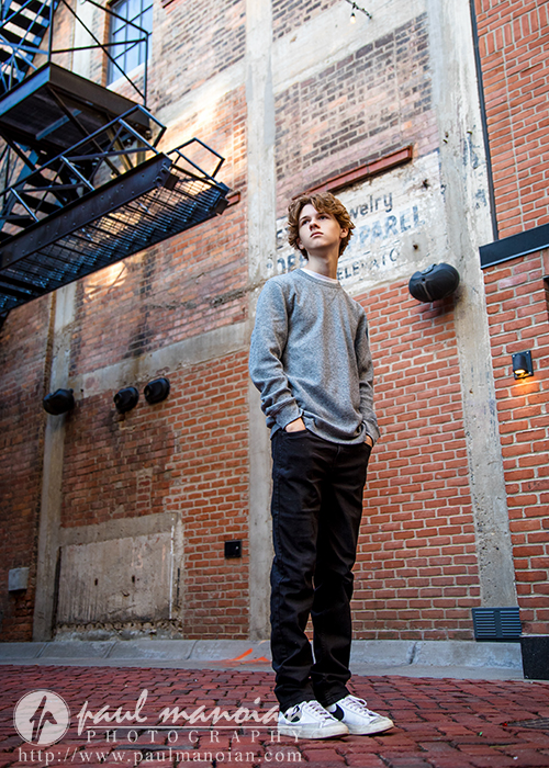A young person in a gray sweater and black pants stands with hands in pockets in an urban alleyway, surrounded by brick walls, a fire escape, and faded signage—capturing the vibe of a Detroit senior pictures session.