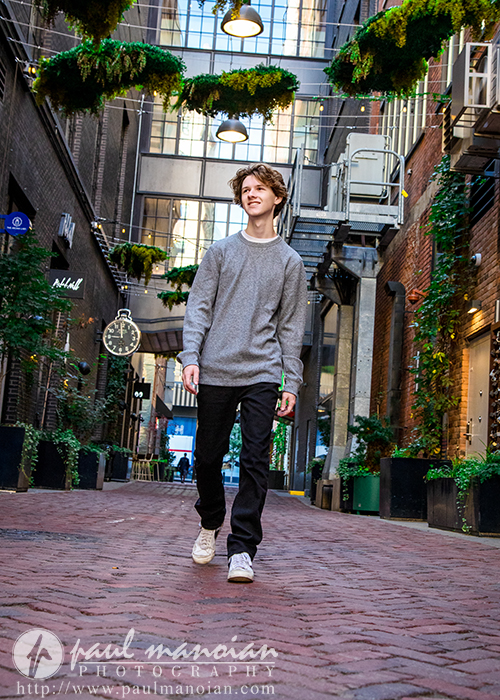 A young man in a gray sweater and black pants walks confidently down a brick alleyway lined with greenery and string lights during his Detroit senior pictures session, with urban buildings and a clock in the background.