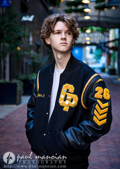 A young person with wavy brown hair wears a black varsity jacket with yellow patches, standing in a brick alley during their Detroit senior pictures session. The jacket displays the number 26 and the name "Tyler S." as they look to the side.