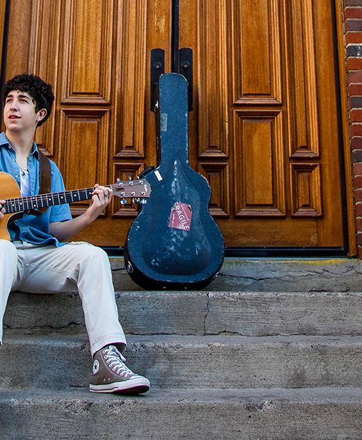A young person sits on outdoor steps in Detroit, playing an acoustic guitar during a session for senior pictures, with a guitar case propped behind them and large wooden doors in the background. They wear a blue shirt, light pants, and gray high-top sneakers.