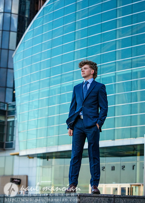 A young man in a blue suit stands confidently outdoors in front of a modern glass building during a session for Northville senior pictures in downtown Detroit, looking to the side with his hands in his pockets.