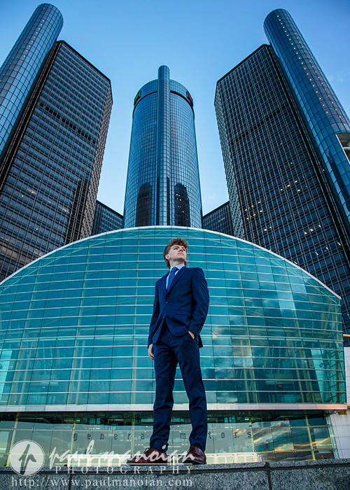 A man in a blue suit stands confidently in front of a modern glass building, as if captured during a session for Northville senior pictures in downtown Detroit, with tall skyscrapers rising behind him against the clear sky.