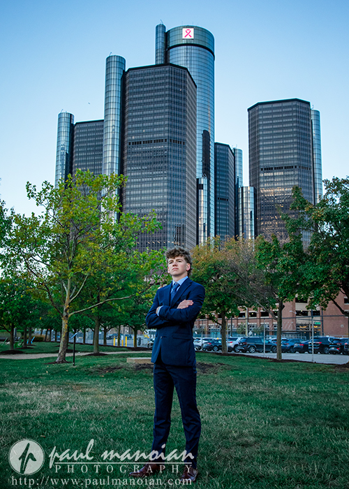 A young man in a blue suit stands with arms crossed on grass, skyscrapers and trees behind him—a session for Northville senior pictures in downtown Detroit. The photo is watermarked "Paul Manoian Photography.