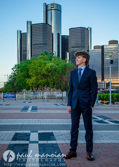 A young man in a navy suit stands outdoors on a patterned plaza, smiling and looking to the side during a session for Northville senior pictures in downtown Detroit, with glass skyscrapers and green trees in the background under a clear sky.