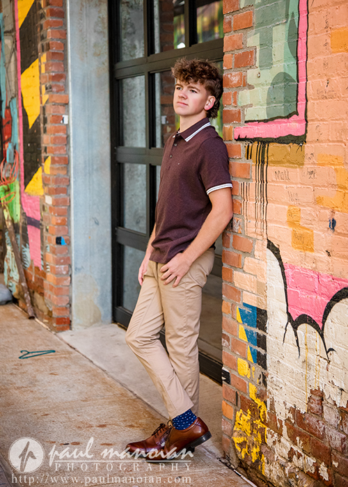 A teenage boy in a maroon polo shirt and beige pants leans against a colorful brick wall with graffiti, posing near a doorway during a session for Northville senior pictures in downtown Detroit. He looks off to the side with one hand in his pocket.