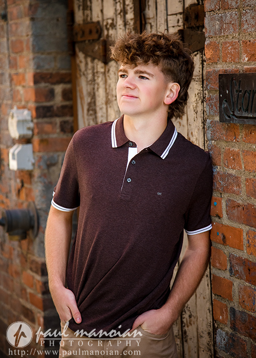 A young person with curly hair, wearing a dark brown polo shirt, stands against a rustic brick wall and wooden door, looking upward with hands in their pockets—a stylish session for Northville senior pictures in downtown Detroit. Paul Manoian Photography logo at the bottom.