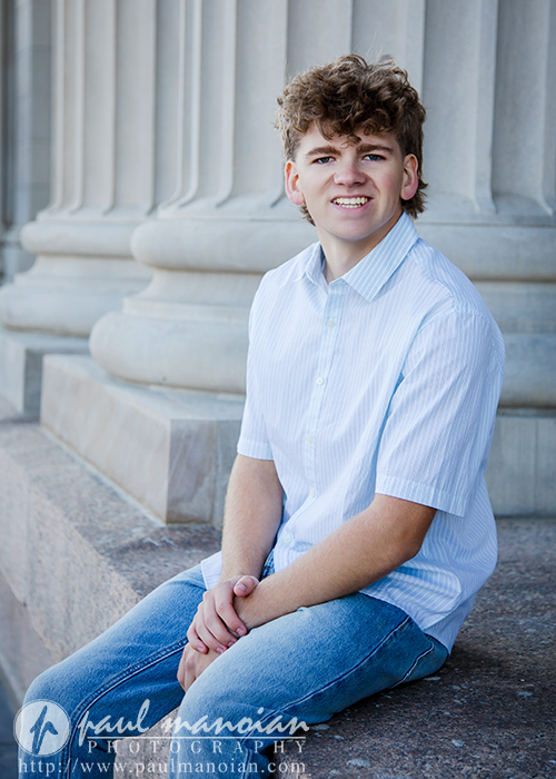 A young man with curly hair, wearing a light blue short-sleeve shirt and jeans, sits on a stone ledge in front of large columns, smiling at the camera during a session for Northville senior pictures in downtown Detroit.