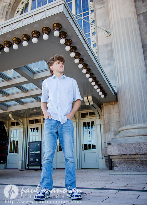 A young person with curly hair, wearing a light blue short-sleeved shirt and jeans, stands confidently outside a grand building with large columns during a session for Northville senior pictures in downtown Detroit.