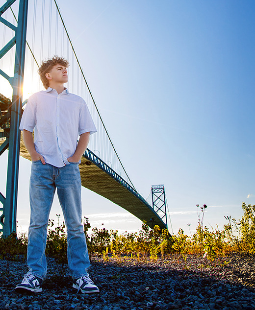 A young person stands on gravel near a tall steel bridge, hands in pockets, looking into the distance during a session for Northville senior pictures in downtown Detroit. Sunlight filters through the structure, with blue sky and scattered plants in the foreground.