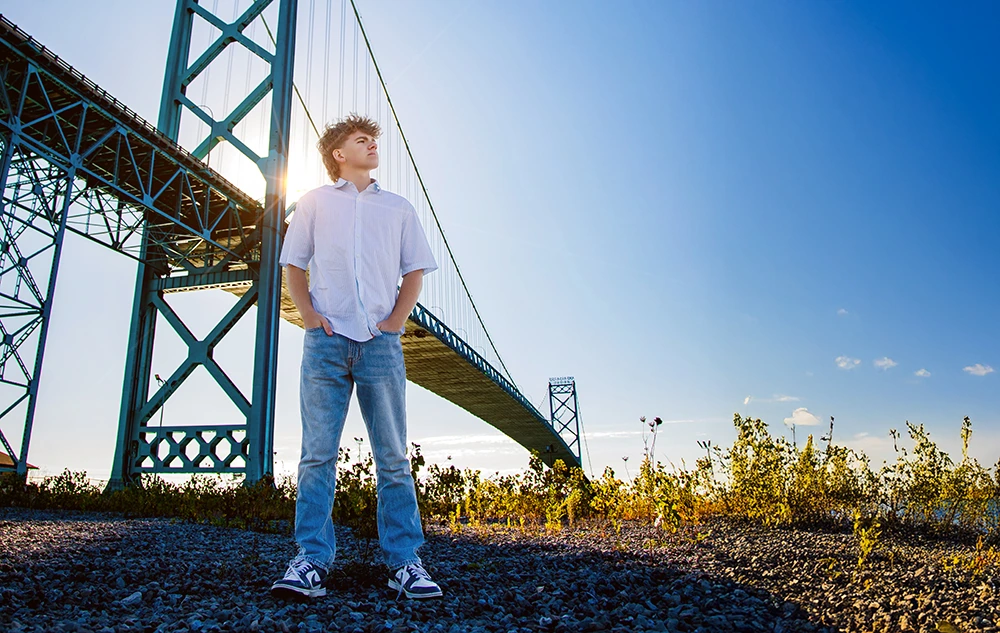 A young person stands on gravel near a tall steel bridge, hands in pockets, looking into the distance during a session for Northville senior pictures in downtown Detroit. Sunlight filters through the structure, with blue sky and scattered plants in the foreground.
