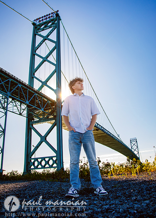 A young man in a white shirt and jeans stands confidently on gravel in front of a large steel bridge, sunlight streaming behind, during a session for Northville senior pictures in downtown Detroit.