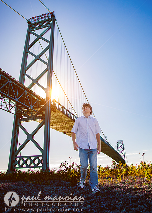 A boy in a white shirt and jeans walks under a large suspension bridge on a sunny day—a perfect session for Northville senior pictures in downtown Detroit, with the sun shining behind the bridge and a clear blue sky above.