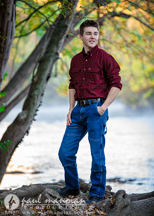 A young man in a red plaid shirt and blue jeans stands confidently on tree roots by a river, surrounded by green trees and soft natural light—an ideal moment from his Ann Arbor senior pictures session.