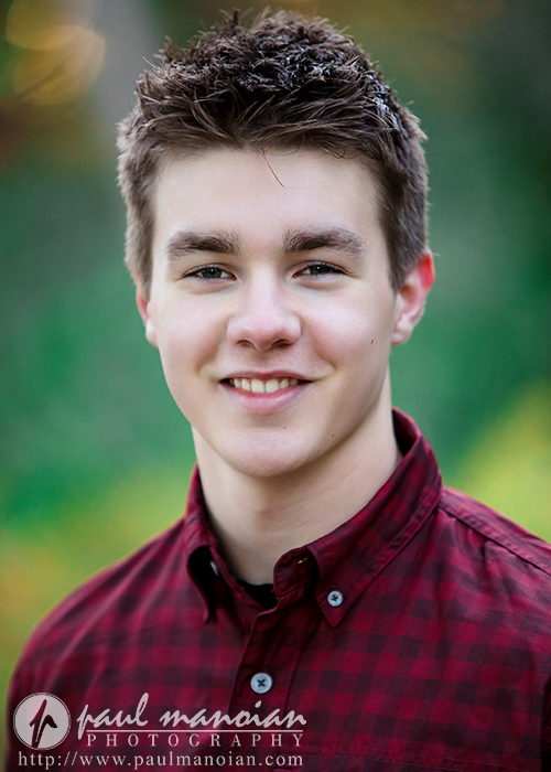 A young man with short brown hair, wearing a red plaid button-up shirt, smiles at the camera outdoors during his Ann Arbor senior pictures session. The logo "paul manoian photography" appears in the lower left corner against a blurred green background.