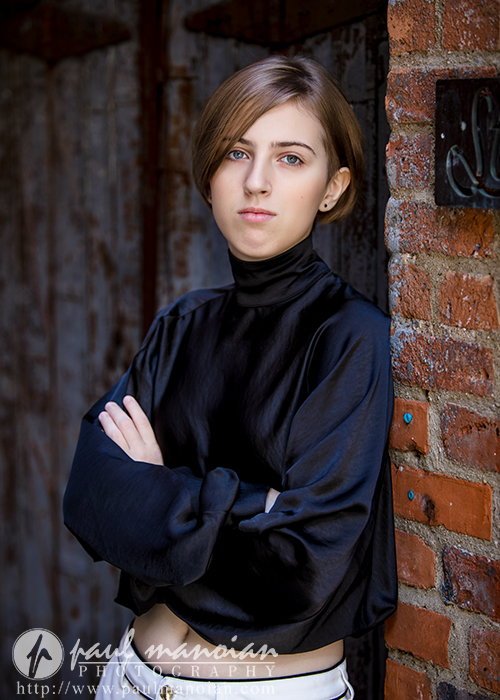 A young person with short light brown hair and a black turtleneck blouse stands with arms crossed, leaning against a brick wall and gazing confidently at the camera during a session for senior pictures in Detroit.