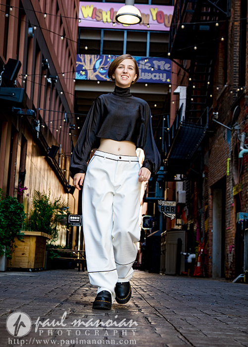 A person wearing a black crop top and white pants walks confidently down a narrow, colorful alleyway decorated with lights and murals during a vibrant session for senior pictures in Detroit. The person is smiling, embracing the lively urban vibe.