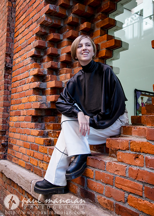 A person with short hair, wearing a black long-sleeve top, white pants, and black platform shoes, sits smiling on a brick window ledge during a session for senior pictures in Detroit’s urban setting.
