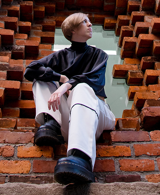A person with short hair, wearing a black top and white pants, sits in a large square opening in a red brick wall—striking a pose for a session for senior pictures in Detroit, looking to the side with one leg raised and resting on the brick ledge.