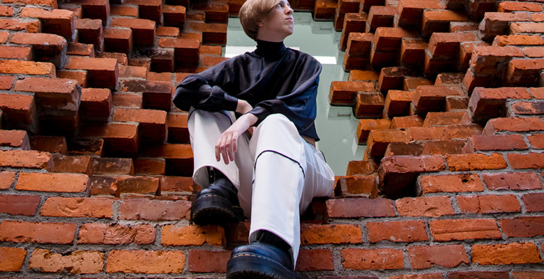 A person with short hair, wearing a black top and white pants, sits in a large square opening in a red brick wall—striking a pose for a session for senior pictures in Detroit, looking to the side with one leg raised and resting on the brick ledge.