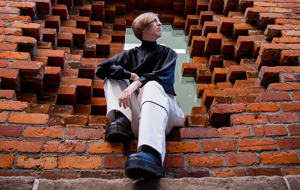 A person with short hair, wearing a black top and white pants, sits in a large square opening in a red brick wall—striking a pose for a session for senior pictures in Detroit, looking to the side with one leg raised and resting on the brick ledge.