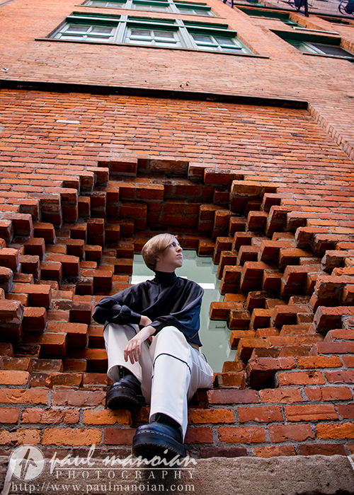 A person with short hair sits in a circular brick window of a tall red brick building, looking up thoughtfully during a session for senior pictures in Detroit. The perspective shows green-trimmed windows stretching high above.