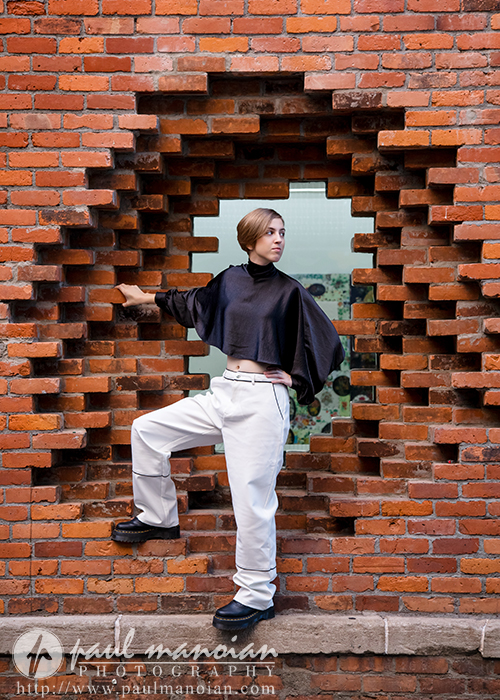 A person poses confidently in front of a unique brick wall with a geometric opening during a session for senior pictures in Detroit, one arm raised and one leg bent, wearing white pants, a black cropped top, and black shoes. Greenery peeks through the opening.