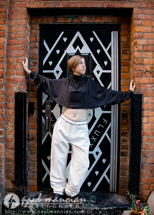 A person wearing a loose black top and white pants poses with arms outstretched against a brick wall, standing in front of a black and white geometric-patterned door labeled "FLUX" during a session for senior pictures in Detroit.