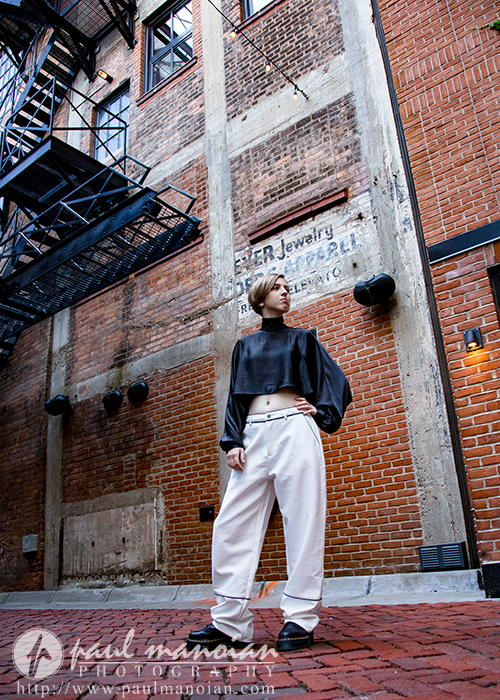 A person in a black long-sleeve crop top and white pants stands confidently in a brick alleyway, with industrial fire escapes and worn vintage signs behind them—perfect for a session for senior pictures in Detroit.