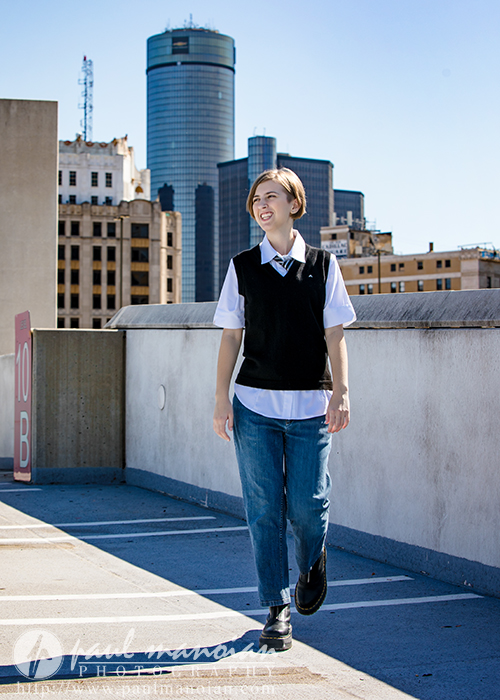 A person in jeans, a white shirt, and a black vest smiles while walking on a rooftop parking lot during a session for senior pictures in Detroit, with tall city buildings in the background under a clear blue sky.