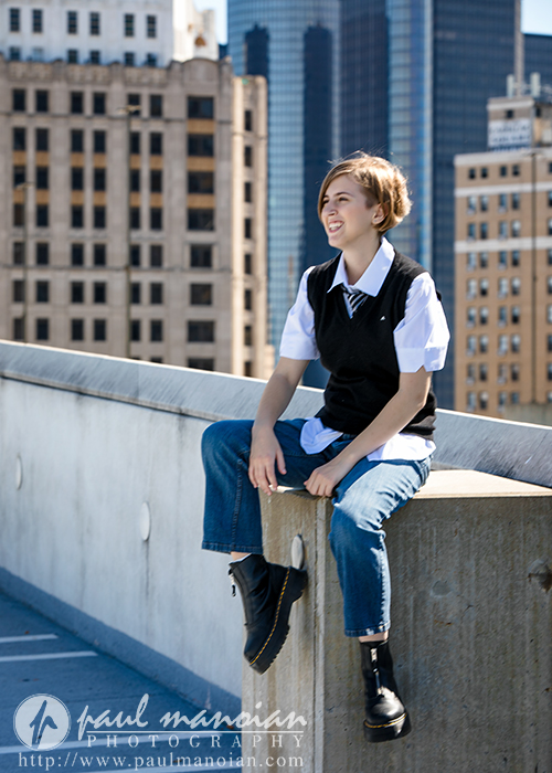 A young person with short hair, dressed in a collared shirt, tie, black vest, and jeans, sits smiling on a concrete ledge during a session for senior pictures in Detroit, with city buildings and skyscrapers in the background.