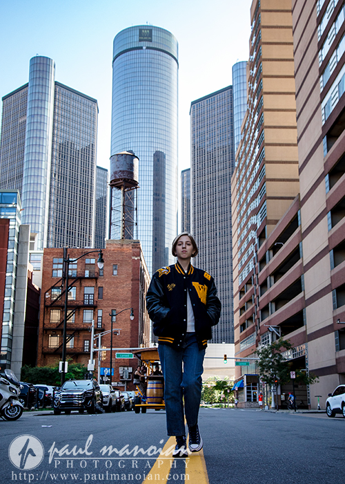 A person in a black and yellow jacket walks confidently down a city street during a session for senior pictures in Detroit, surrounded by tall modern skyscrapers, older brick buildings, and a distant water tower.