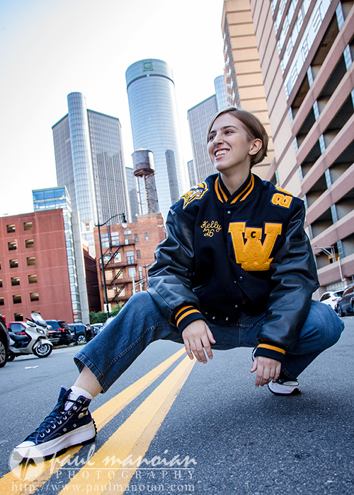 A young woman in a letterman jacket squats and smiles on a city street during her session for senior pictures in Detroit, with tall buildings and skyscrapers in the background. She wears jeans and sneakers, capturing the urban vibe.