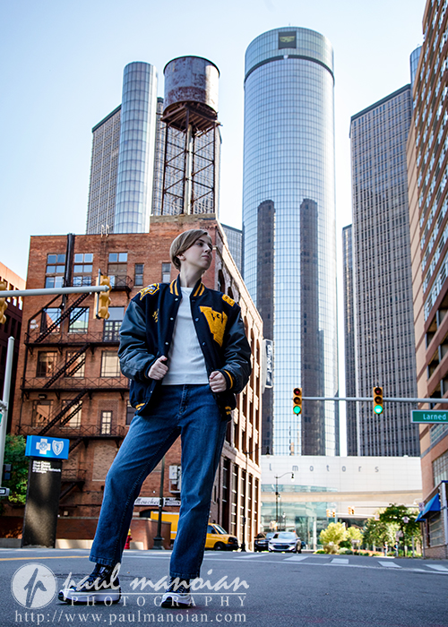 A person wearing a black and yellow varsity jacket and jeans stands confidently in the middle of a city street during a session for senior pictures in Detroit, with tall glass skyscrapers and a water tower visible in the background.
