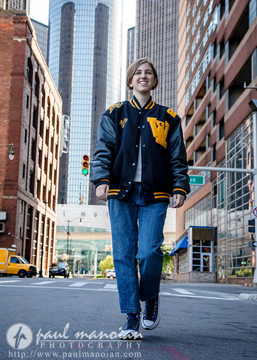 A young person wearing a black and yellow varsity jacket, jeans, and sneakers walks confidently down a city street during a session for senior pictures in Detroit, surrounded by tall buildings and bright daylight.