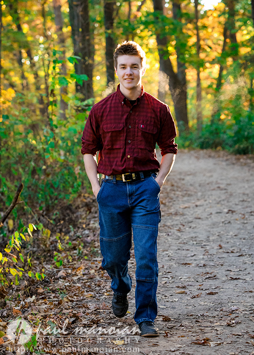 A young man in a red plaid shirt and blue jeans walks along a wooded path with hands in his pockets, smiling, surrounded by green and yellow trees in warm sunlight during his Ann Arbor senior pictures session.
