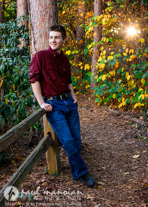 A young man in a maroon shirt and jeans leans against a wooden fence on a forest path during his Ann Arbor senior pictures session, smiling and looking to the side as sunlight streams through autumn trees in the background.