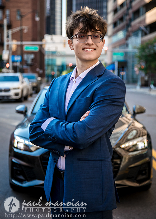 A young man in a blue suit stands confidently with arms crossed on a city street during his Detroit senior pictures session, smiling. A black car is parked behind him, and tall buildings surround the scene. Photography credit appears at the bottom.