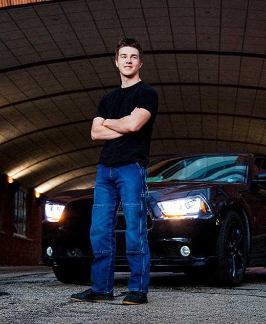 A young man in a black T-shirt and blue jeans stands with arms crossed in front of a black car with headlights on, under an arched brick ceiling—capturing the perfect Ann Arbor senior pictures session vibe in an urban setting.