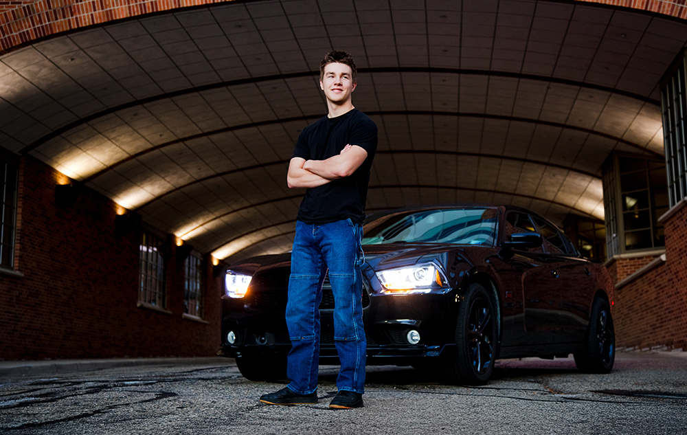 A young man in a black T-shirt and blue jeans stands with arms crossed in front of a black car with headlights on, under an arched brick ceiling—capturing the perfect Ann Arbor senior pictures session vibe in an urban setting.