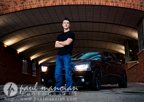 A young man in a black t-shirt and blue jeans stands confidently with arms crossed in front of a black car under a brick archway, its headlights on—captured during an Ann Arbor senior pictures session. Photography watermark is visible at the bottom.