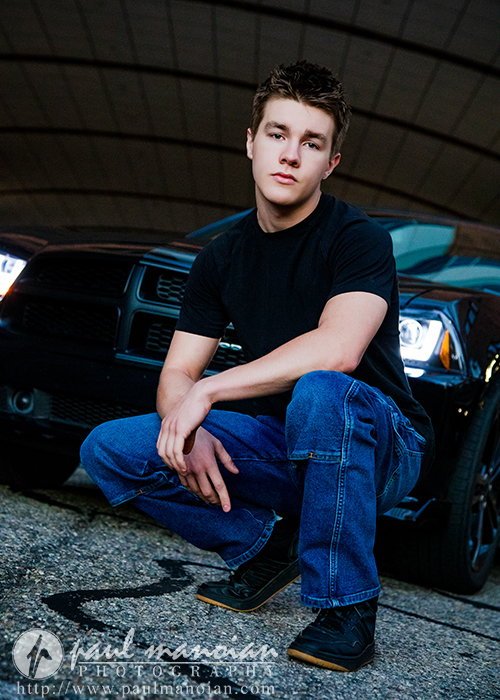 A young man in a black t-shirt and jeans crouches in front of a black car, looking at the camera during his Ann Arbor senior pictures session. The scene is set outdoors beneath a large, curved structure.