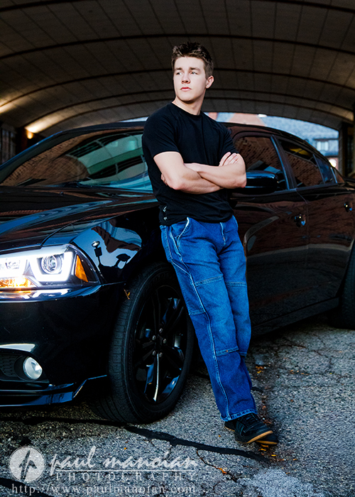 A young man in a black t-shirt and blue jeans leans against a shiny black car parked under an overpass, looking off to the side with his arms crossed during his Ann Arbor senior pictures session.