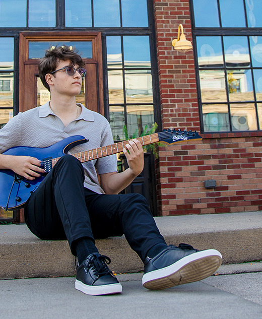 A young person with glasses sits on a concrete step outside a brick building, playing a blue electric guitar. They wear a light polo shirt, black pants, and sneakers. Reflections are visible in the large windows behind them.
