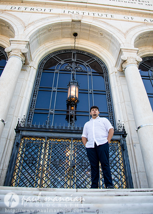 A man in a white shirt stands on the steps in front of the grand entrance of the Detroit Institute of Arts, perfectly capturing a Detroit senior portraits session amid tall columns, ornate iron gates, and a large hanging lantern above him.