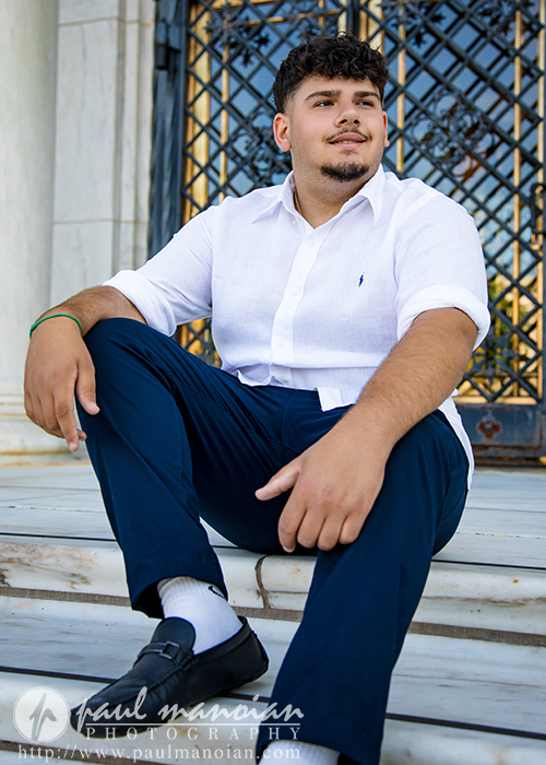 A young man with short dark hair and a beard sits on marble steps during his Detroit senior portraits session, wearing a white button-up shirt and navy pants. He looks to the side with a slight smile, ornate metalwork and a white column behind him.