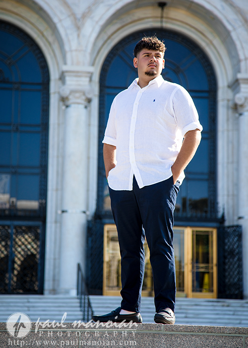 A young man in a white button-up shirt and dark pants stands confidently on stone steps outside a large building with arched windows and ornate architecture during his Detroit senior portraits session.