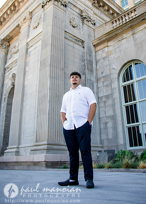 A young man in a white shirt and dark pants stands confidently in front of a grand, historic stone building with tall columns and arched windows during his Detroit senior portraits session.