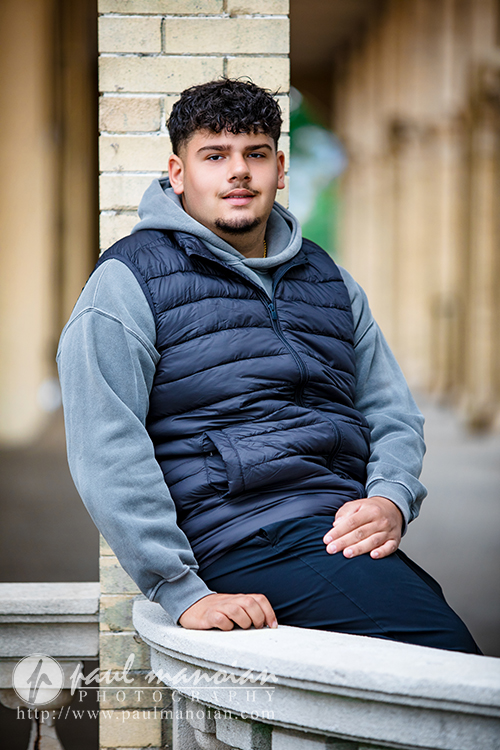 A young man with curly hair, wearing a gray hoodie and black puffer vest, sits casually against a brick wall outdoors, looking at the camera with a relaxed expression during his Detroit senior portraits session.